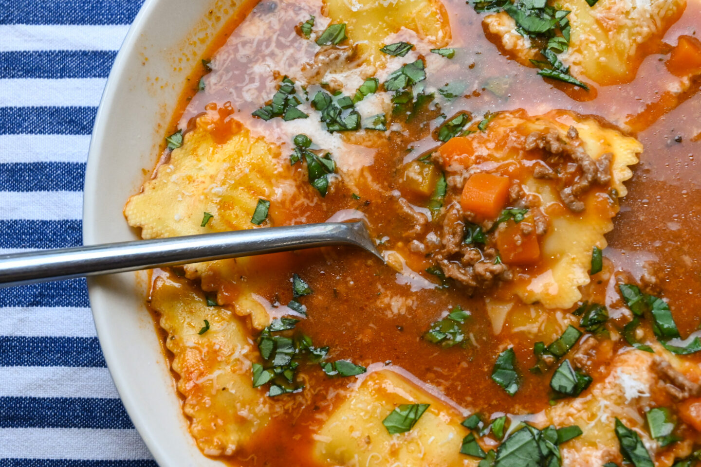 Bowl of soup featuring cheese ravioli, ground beef and cubed carrots in a tomato red broth sprinkled with Parmesan cheese and chopped fresh basil