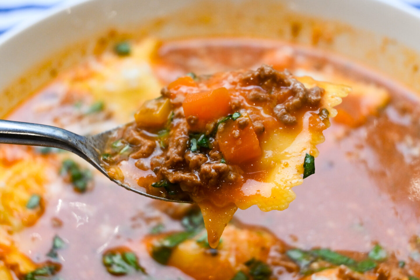 closeup of a spoonful of soup featuring cheese ravioli, ground beef and cubed carrots in a tomato red broth sprinkled with Parmesan cheese and chopped fresh basil 