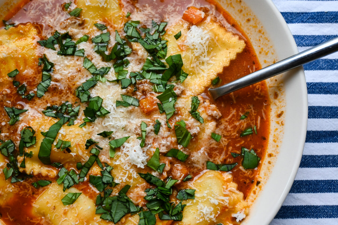 Bowl of soup featuring cheese ravioli, ground beef and cubed carrots in a tomato red broth sprinkled with Parmesan cheese and chopped fresh basil