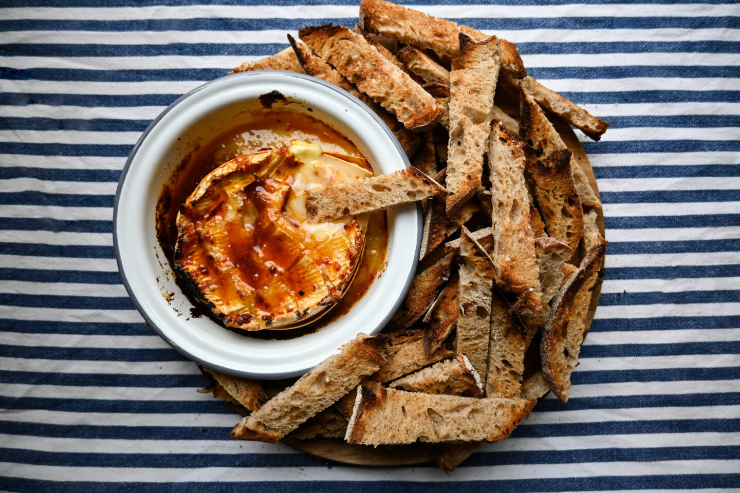 Harissa Honey Baked Camembert With Toast Points on a round serving board with a blue & white striped background